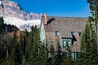 Une cabane en bois aux volets verts, entourée d’une forêt dense de conifères, avec un glacier et des montagnes en toile de fond.