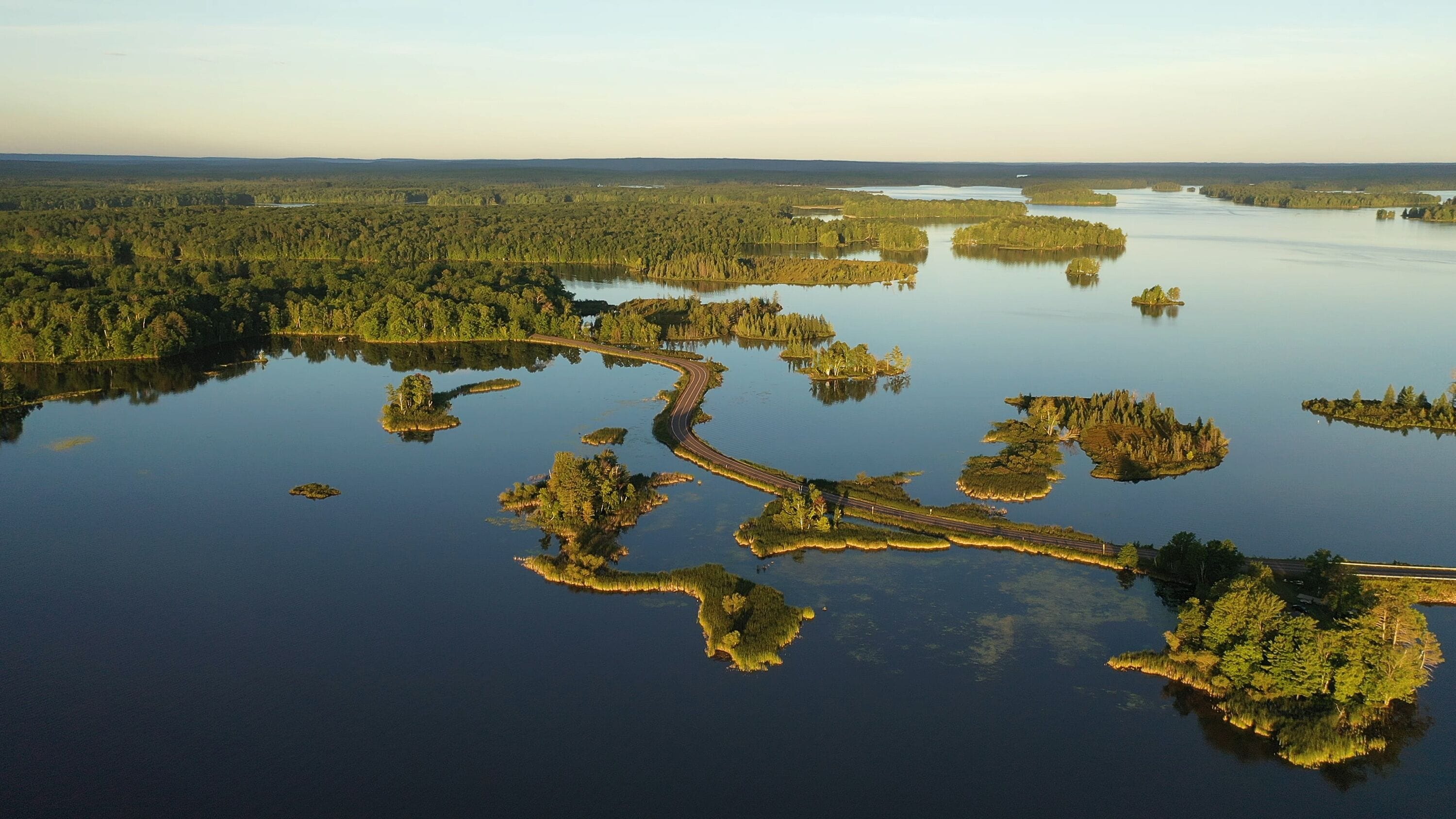 Establishing shot of american wilderness. Aerial view of scenic road through lake, and wooded islands. Calm morning water, dawn sky, view from above.