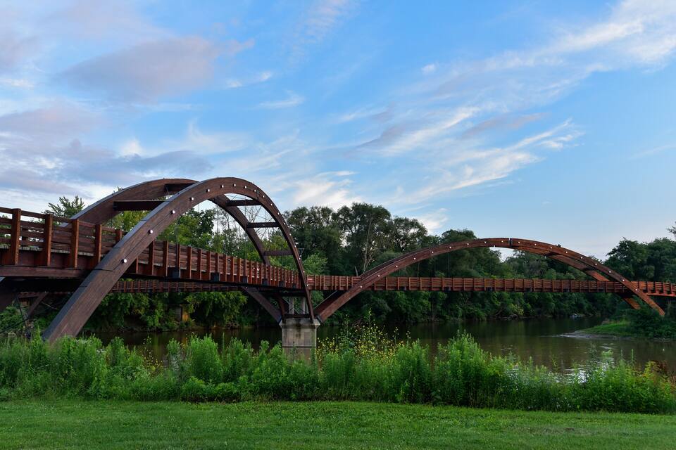 A metal bridge with a curved design over a river, surrounded by greenery and trees.
