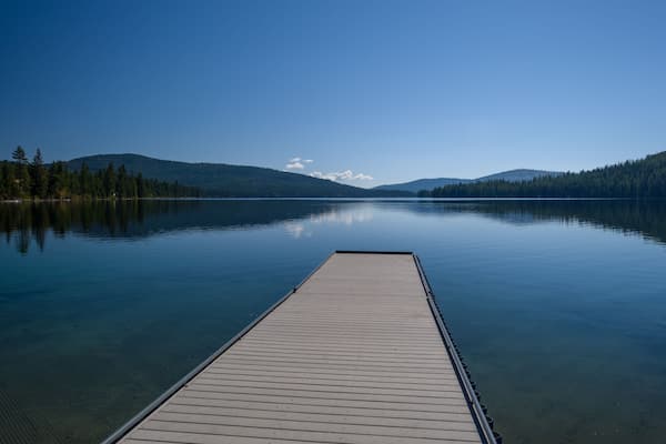 A calm lake with a wooden dock extending into the water, surrounded by forested hills under a clear blue sky.