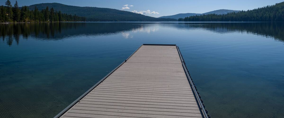 A calm lake with a wooden dock extending into the water, surrounded by forested hills under a clear blue sky.