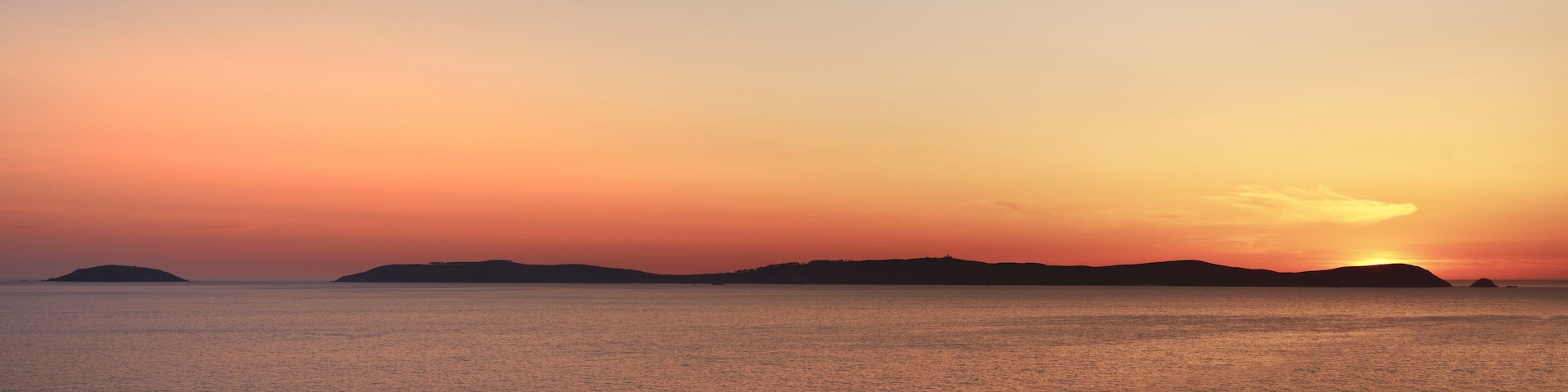 Ein Sonnenuntergang über einem ruhigen Meer, im Hintergrund schattenhafte Inseln am Himmel.