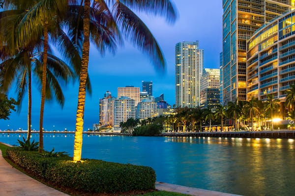 Paisagem urbana à beira-mar com palmeiras, um rio e edifícios altos ao entardecer.