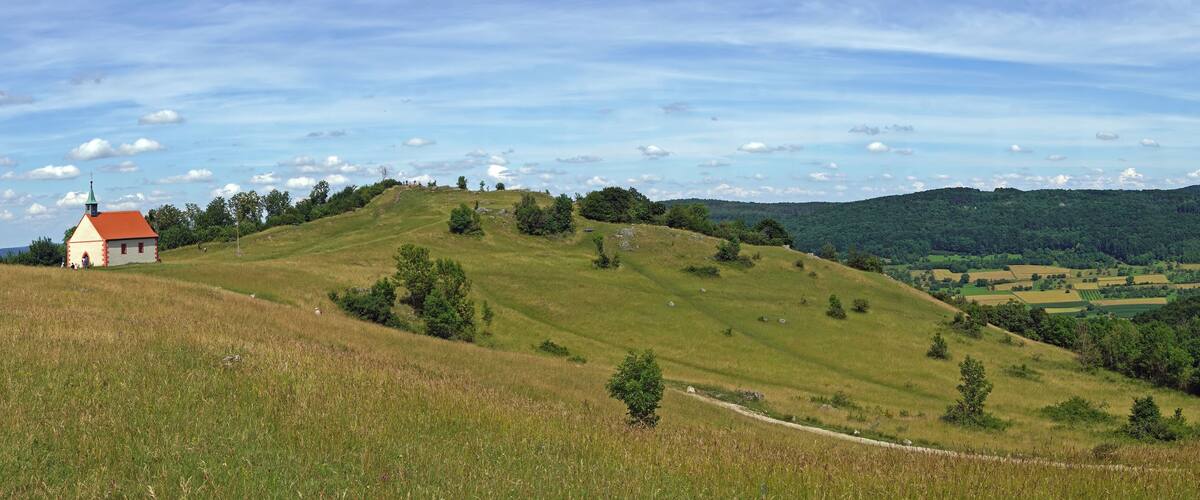 A small church on a grassy hill with a red roof, surrounded by lush green hills and a clear sky.