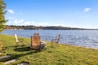 A lakeside view with two chairs and a fire pit on a grassy area.