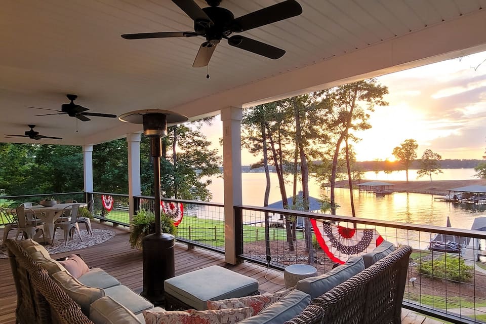 A covered deck with outdoor furniture, ceiling fans, and a view of a lake with a dock and buildings in the distance.