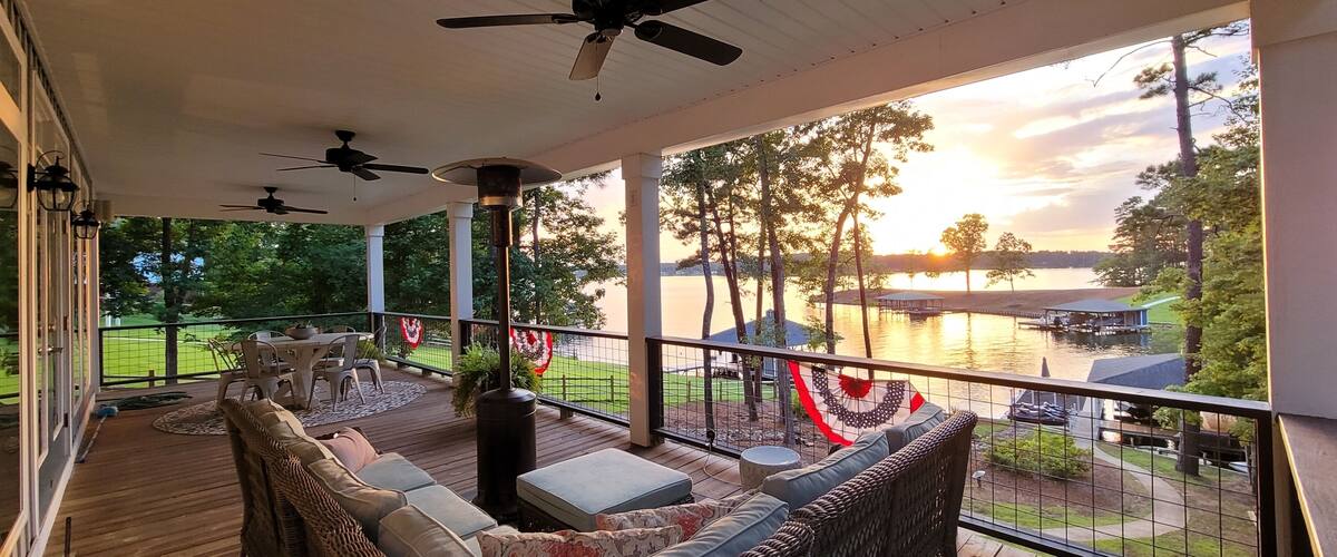 A covered deck with outdoor furniture, ceiling fans, and a view of a lake with a dock and buildings in the distance.