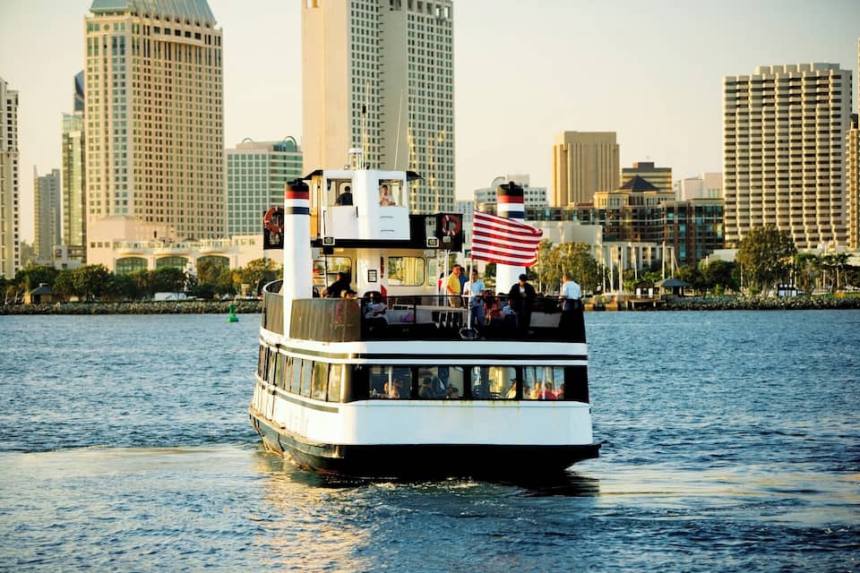 A riverboat with an American flag, passengers, and a city skyline in the background.