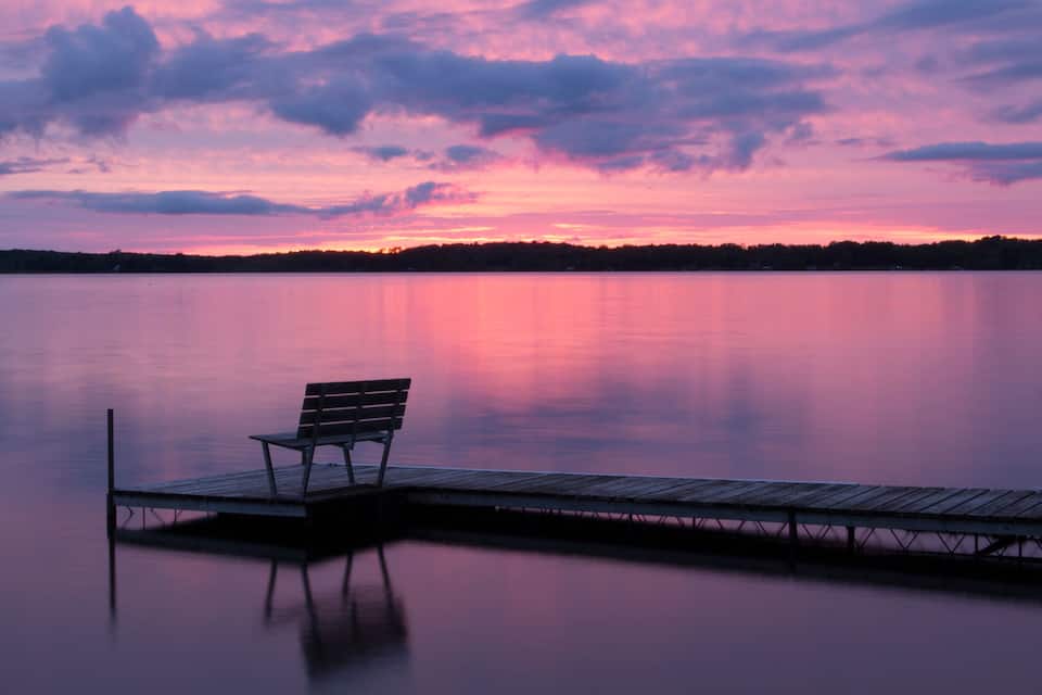 A wooden dock extending into a calm lake with a bench on it, under a colorful sunset sky.