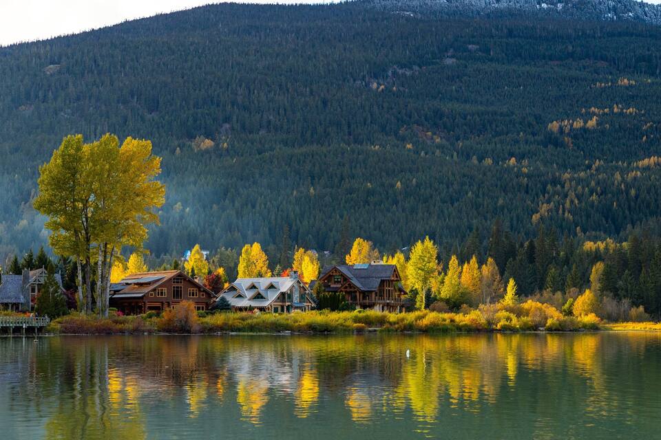 Un conjunt de cabanes al costat d'un llac, amb tardor i un paisatge de muntanya boscosa de fons.