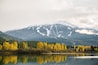 A mountain range with snow-capped peaks, a lake reflecting the trees, and a forest with autumn-colored foliage.