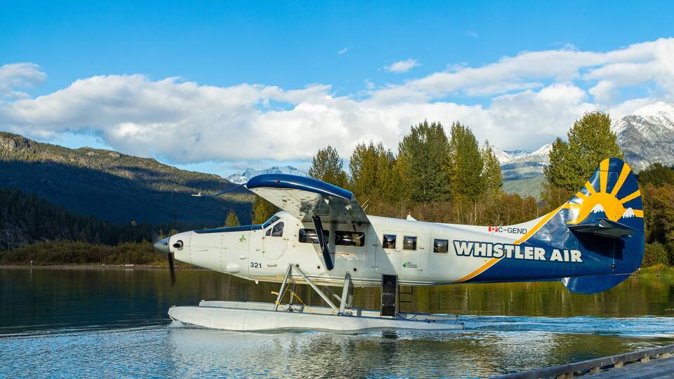 A seaplane with the text "Whistler Air" on its side, docked at a lake with mountains in the background.