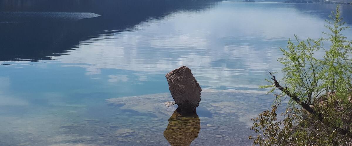 A calm lake with a prominent rock in the foreground and mountains in the background.