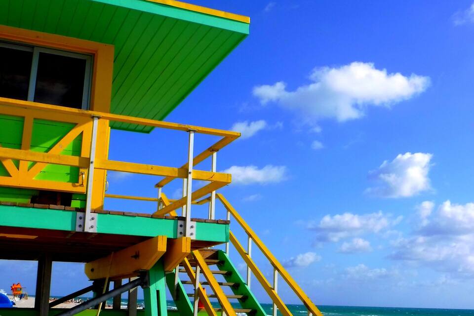 A lifeguard tower with green and yellow colors, stairs, and a beach in the background.