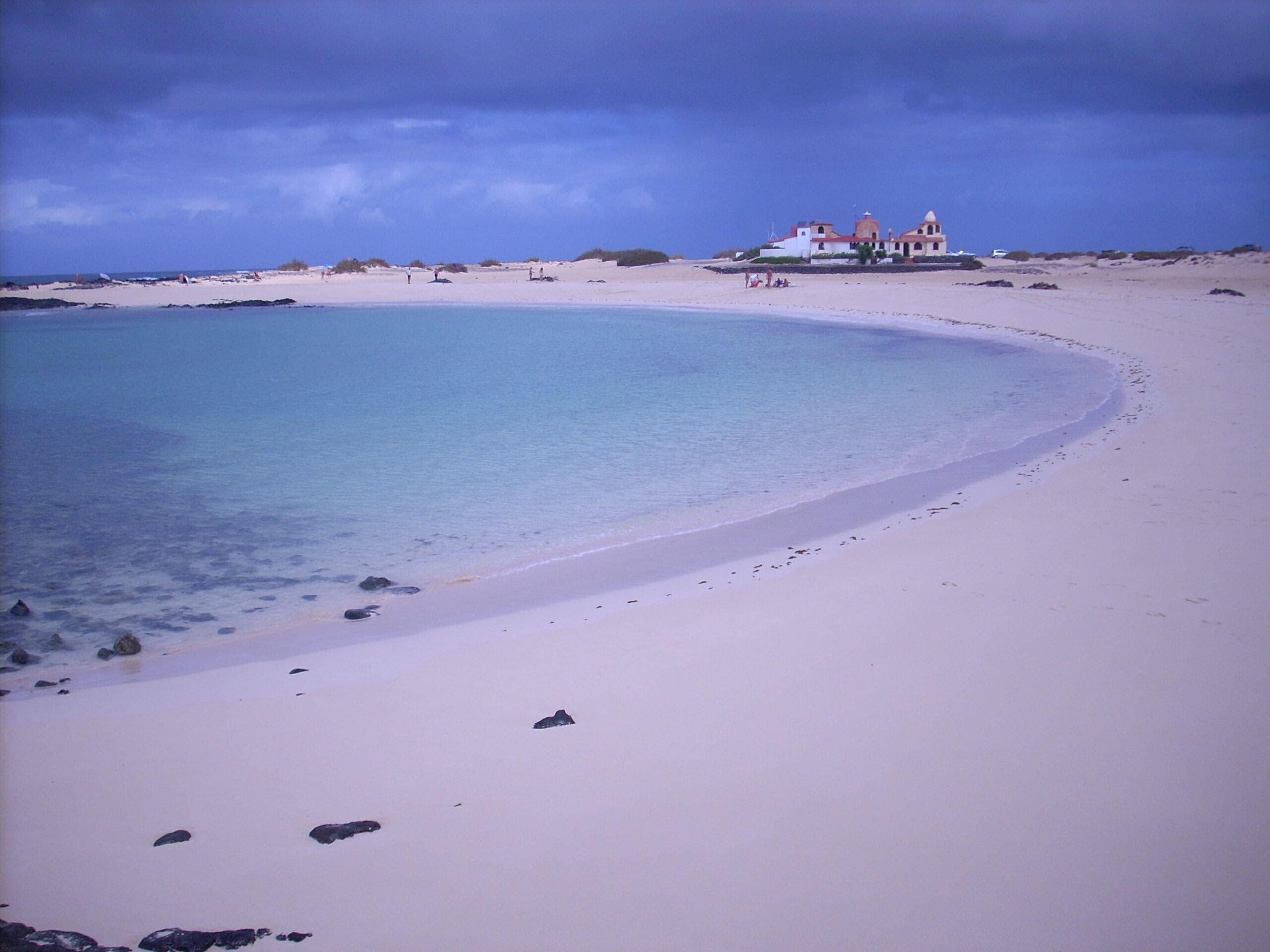 Una playa con aguas turquesas cristalinas, arena blanca y un edificio de techos rojos al fondo.