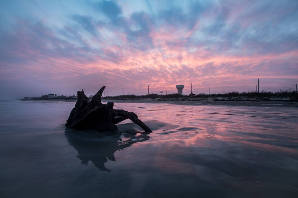 Un coucher de soleil sur une plage, avec une grande formation rocheuse et une tour qui se dresse au loin.