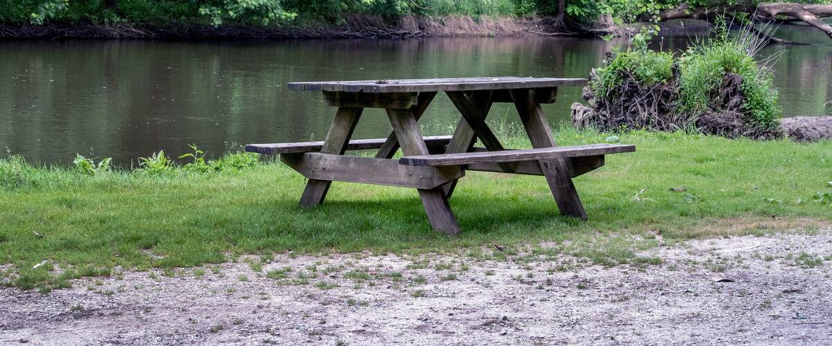 A wooden picnic table by a calm river surrounded by lush greenery.