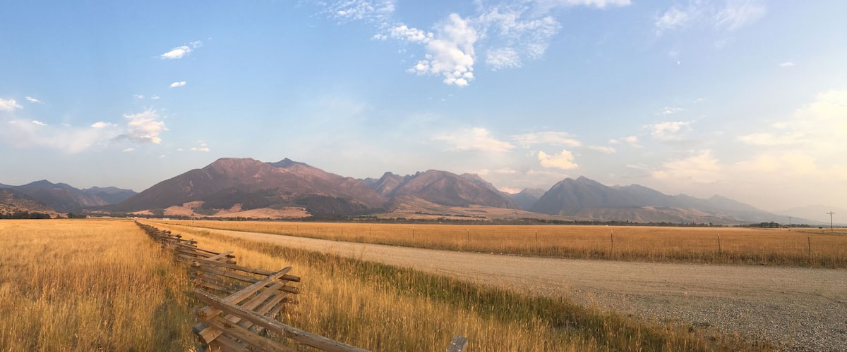 A rural landscape with a dirt road, a wooden fence, and a field of tall grass leading to a range of mountains under a clear sky.