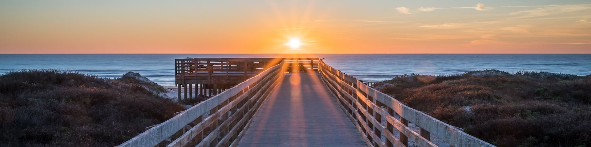 A wooden boardwalk leading to a bench overlooking the ocean at sunset.
