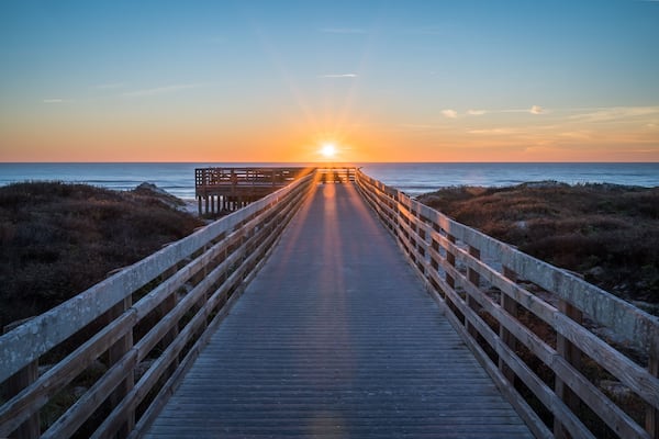 A wooden boardwalk leading to a bench overlooking the ocean at sunset.