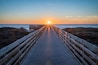 A wooden boardwalk leading to a bench overlooking the ocean at sunset.
