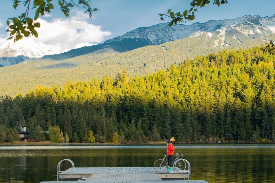 A person in a red jacket standing on a dock by a lake with a forest and mountains in the background.