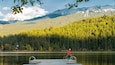 Una persona con una chaqueta roja parada en un muelle junto a un lago, con un bosque y montañas al fondo.