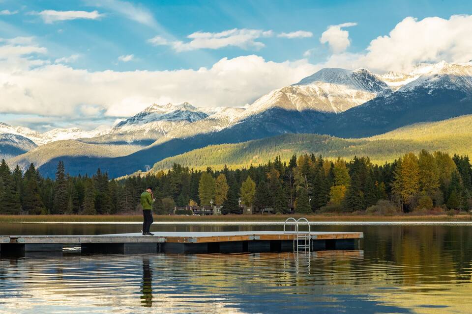 Una persona parada en un muelle junto a un lago tranquilo, con montañas nevadas al fondo.