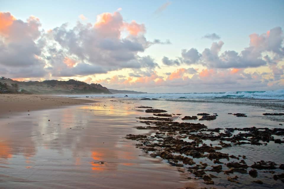 A beach with wet sand and scattered rocks, a cloudy sky, and distant hills.