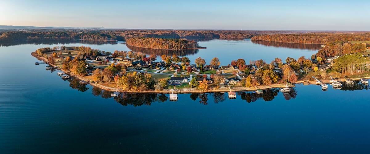 Ett flygbildsbild av en bebyggelse vid en sjö, med hus, bryggor och höstfärgat lövverk.