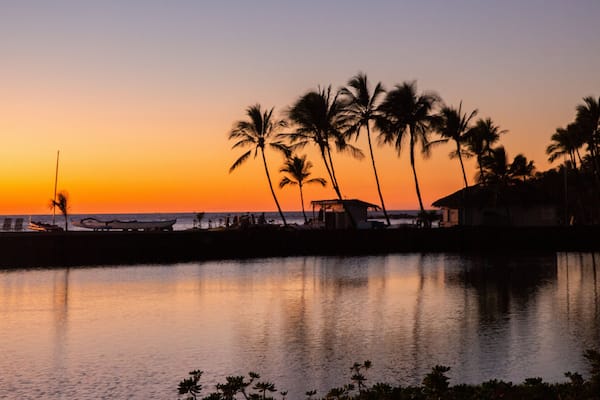A sunset over a calm body of water with palm trees and a beach in the background.