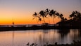 Un coucher de soleil sur une étendue d'eau calme, avec des palmiers et une plage en arrière-plan.