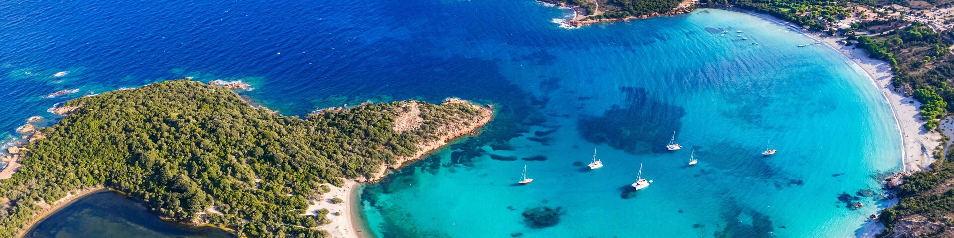 A coastal landscape with clear turquoise waters, a sandy beach, and several sailboats anchored near the shore.