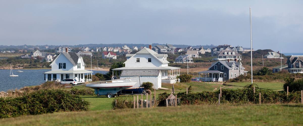 A coastal town with white houses, a boat docked on the shore, and a view of the sea with boats in the distance.
