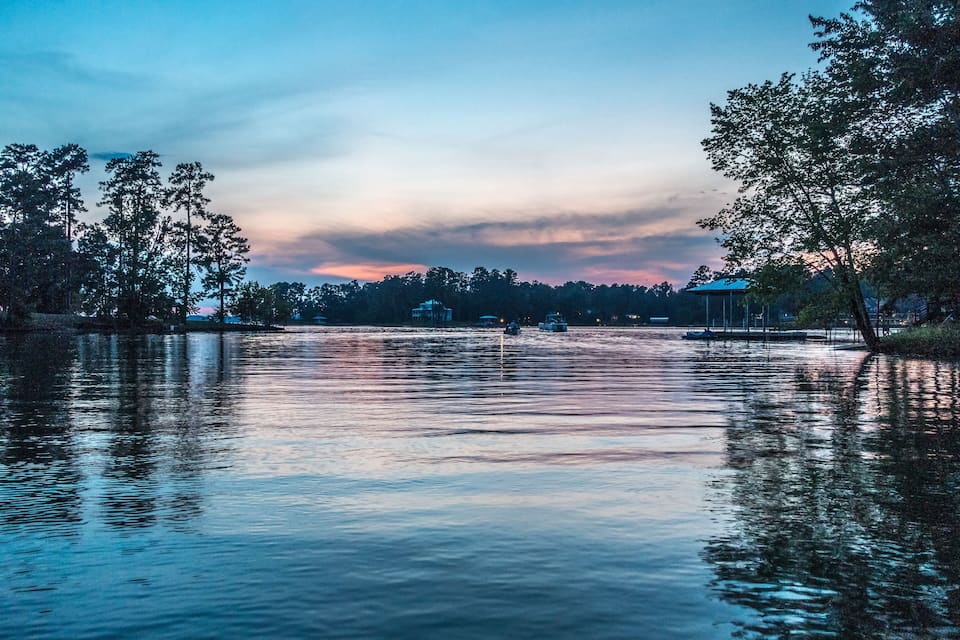 A calm lake at dusk with reflections of trees and a distant building.
