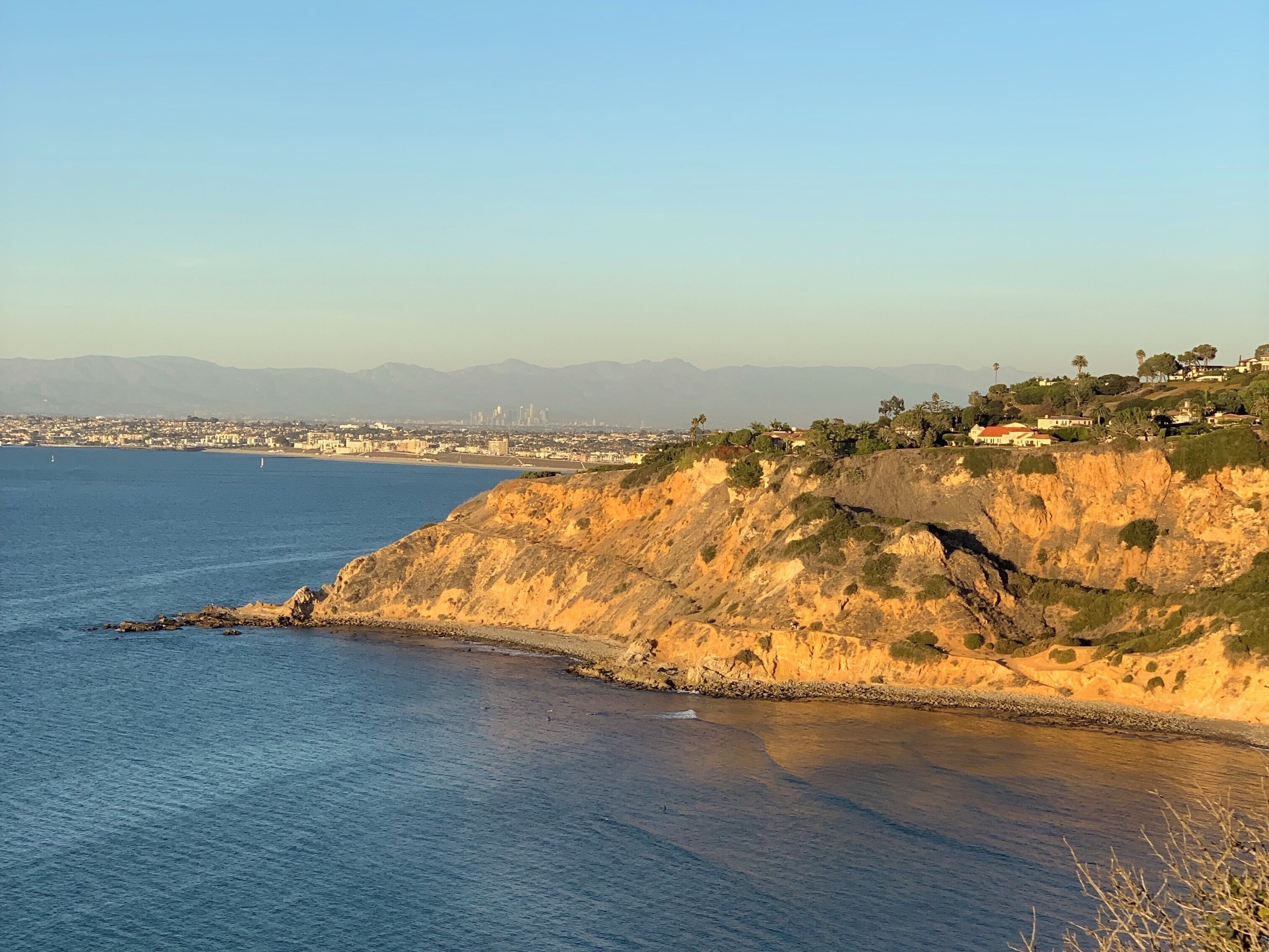 A coastal landscape with a clear view of the ocean, a rocky cliff, and a distant city skyline.