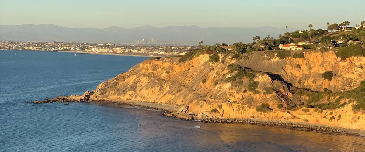 A coastal landscape with a clear view of the ocean, a rocky cliff, and a distant city skyline.