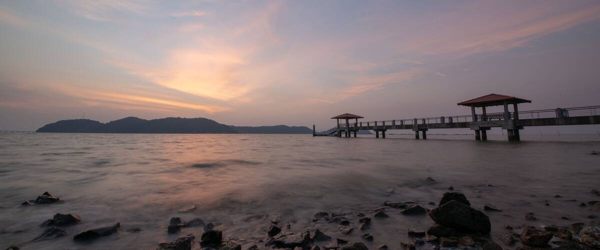 A pier extending into calm waters with a rocky foreground and distant mountains under a pastel sky.