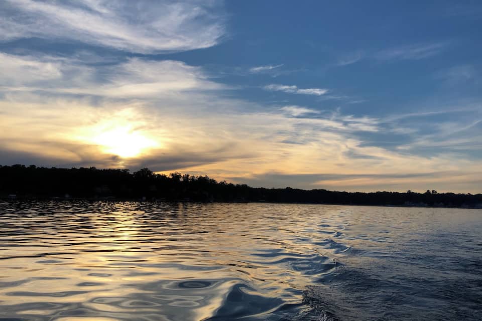 Un coucher de soleil sur un lac paisible, avec de légères ondulations à la surface de l’eau et la silhouette d’arbres en arrière-plan.