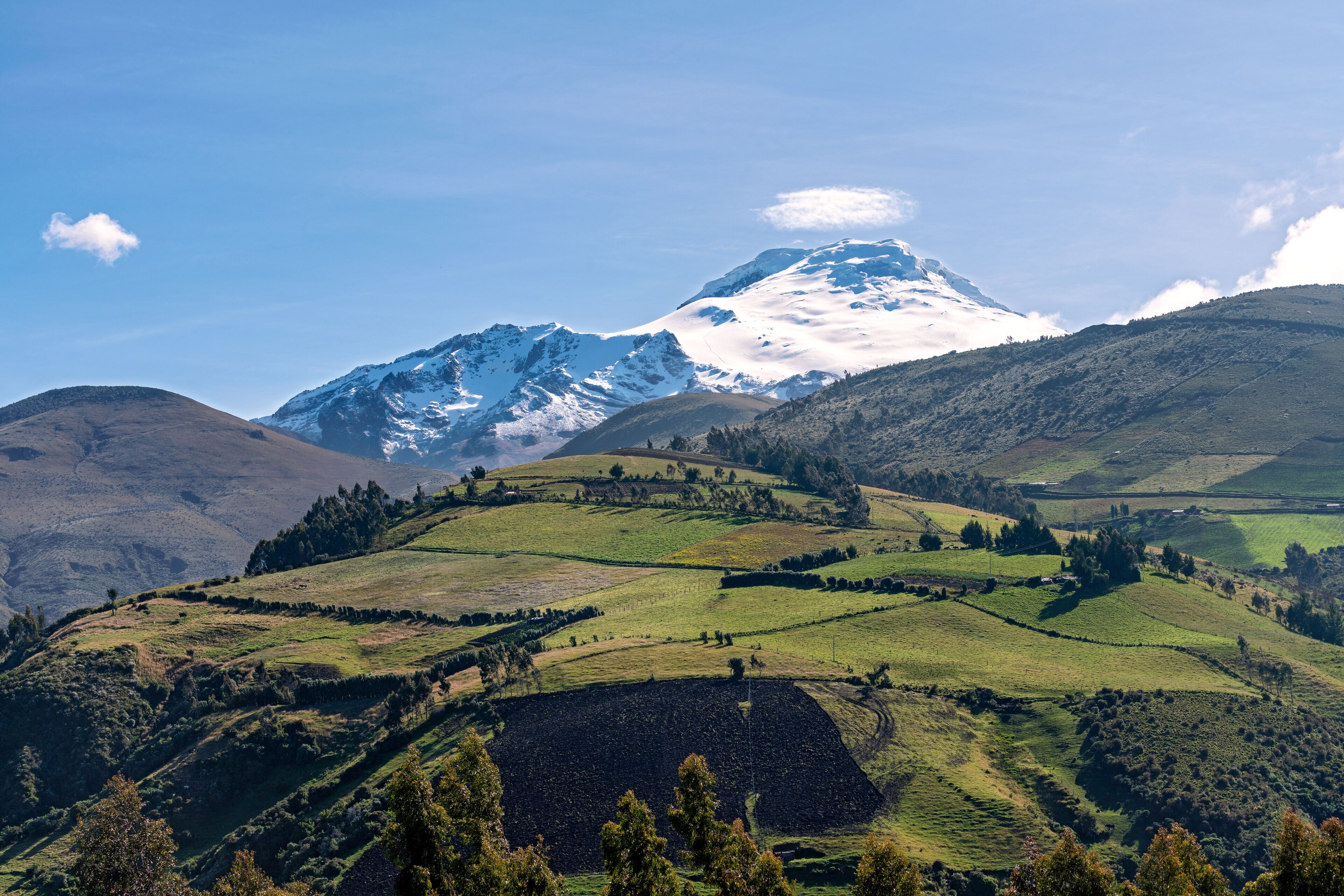 A snow-capped mountain, green rolling hills, and a clear blue sky.