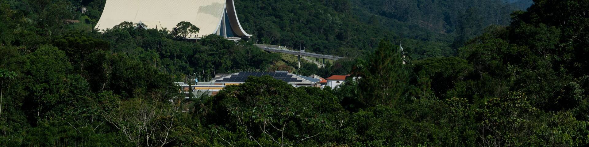 Un edificio grande y blanco con un diseño de techo único, rodeado de un bosque verde denso y colinas al fondo.