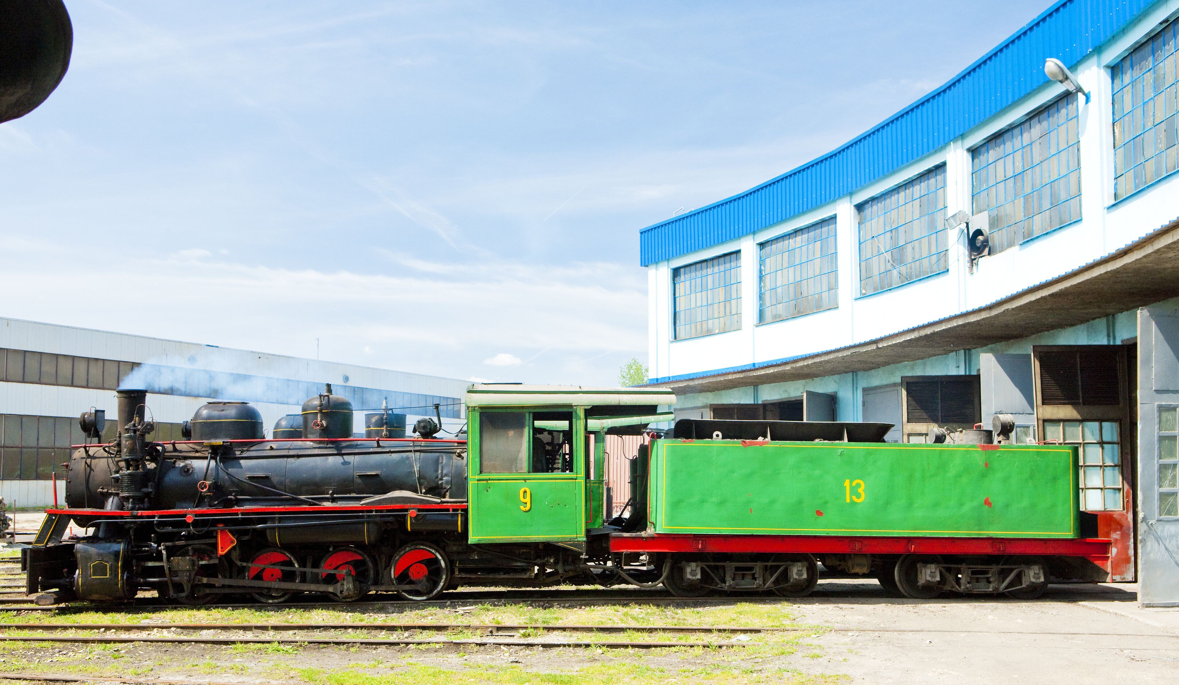 A steam locomotive with a green tender car parked in front of a building with blue and white walls.