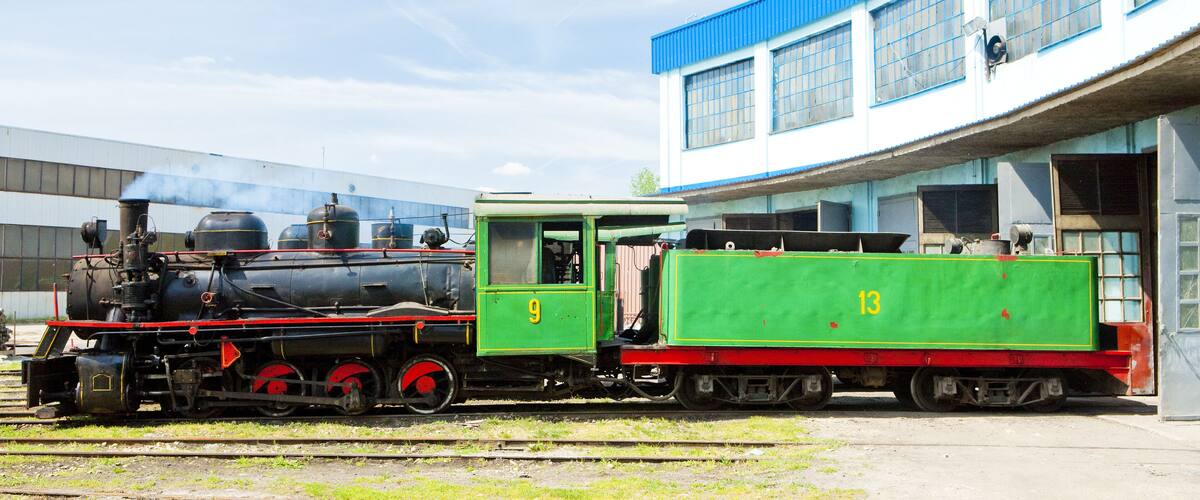 A steam locomotive with a green tender car parked in front of a building with blue and white walls.