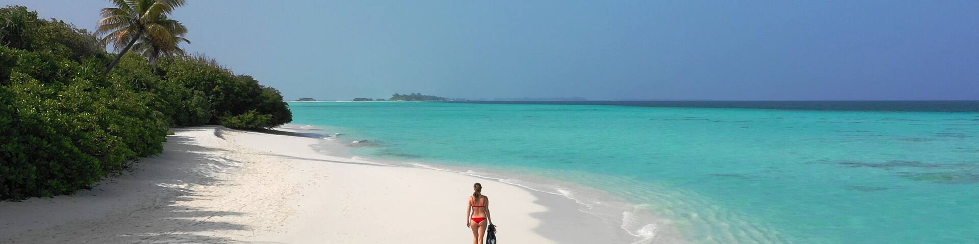 Young woman in a red bikini holding snorkeling gear while walking down a white sand beach. Dhigurah island, Maldives.