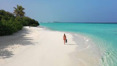 Young woman in a red bikini holding snorkeling gear while walking down a white sand beach. Dhigurah island, Maldives.