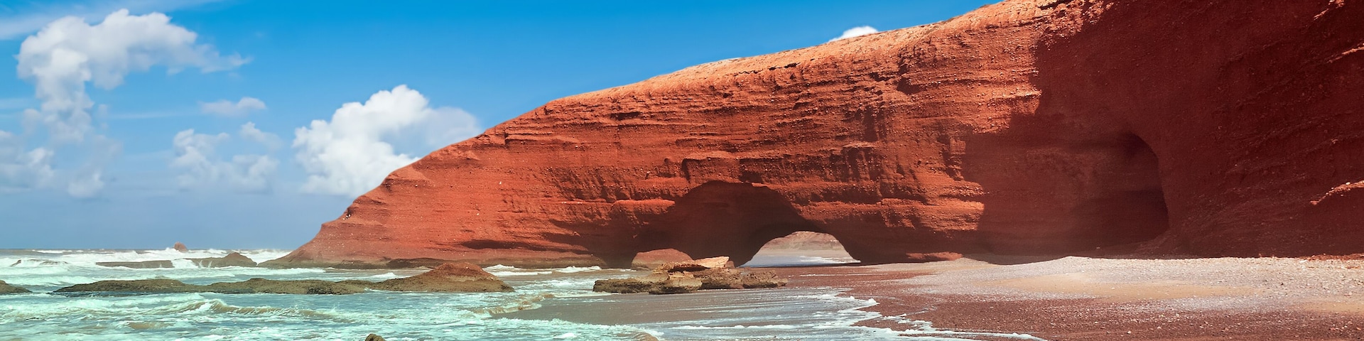 Un paisaje costero con una destacada formación rocosa roja y un cielo azul despejado.
