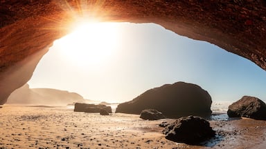 A beach with a large rock arch and the sun shining through it.