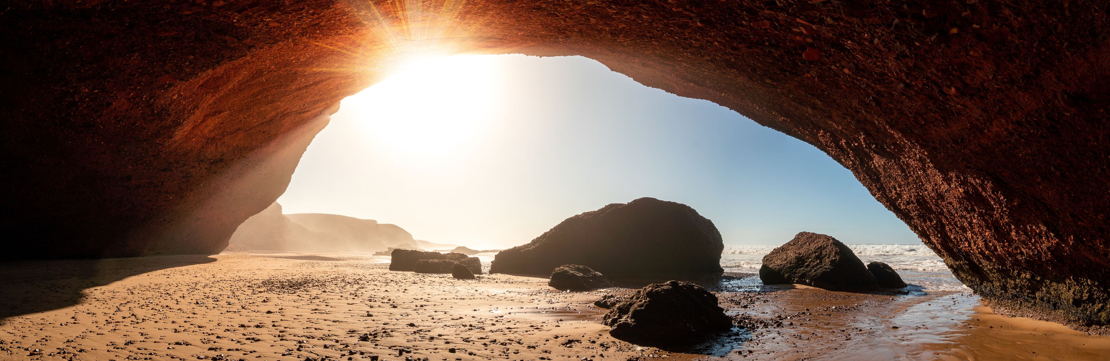 A beach with a large rock arch and the sun shining through it.
