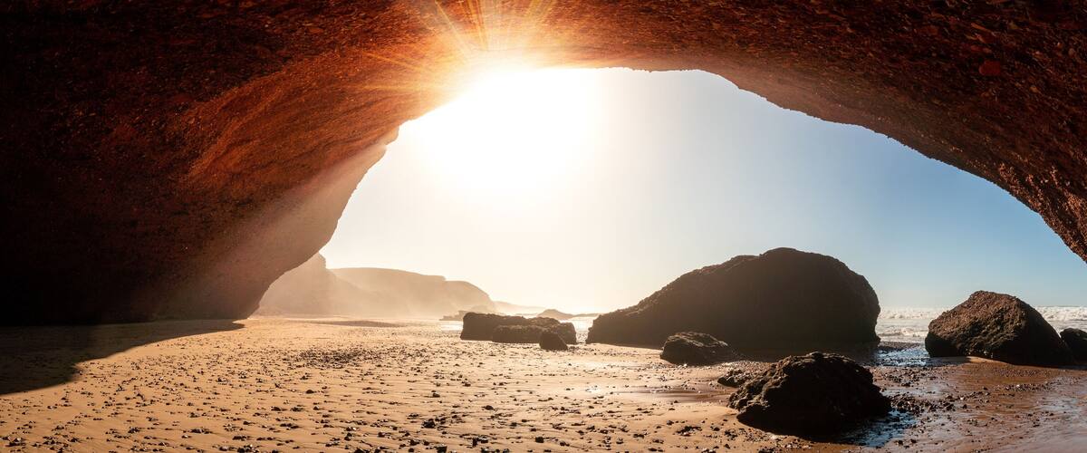 A beach with a large rock arch and the sun shining through it.