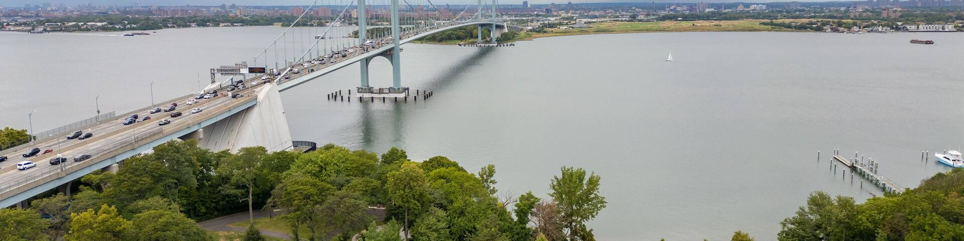 A scenic view of the Whitestone Bridge crossing the East River, connecting urban and natural landscapes. The nearby park offers green spaces with river views, with the horizon and the bustling city of